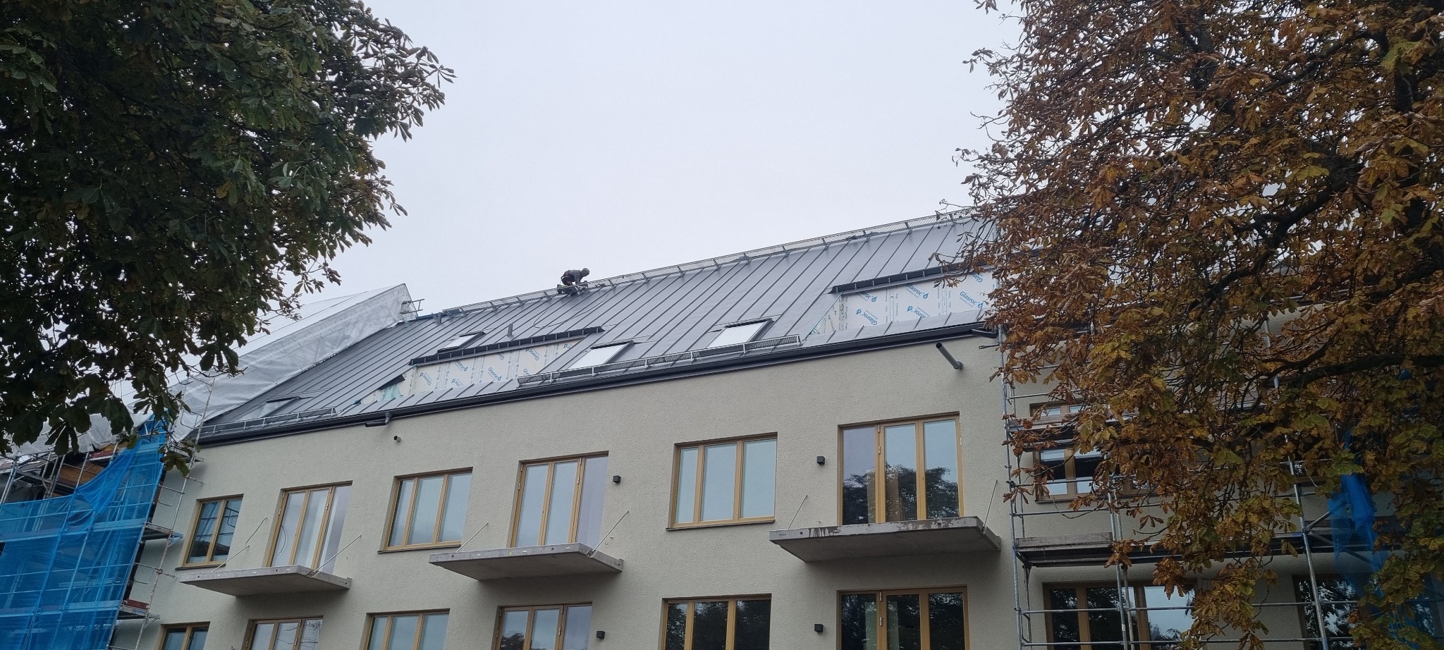 Pracownik na dachu budynku wielorodzinnego z metalowym pokryciem, widoczne okna dachowe i rusztowania.