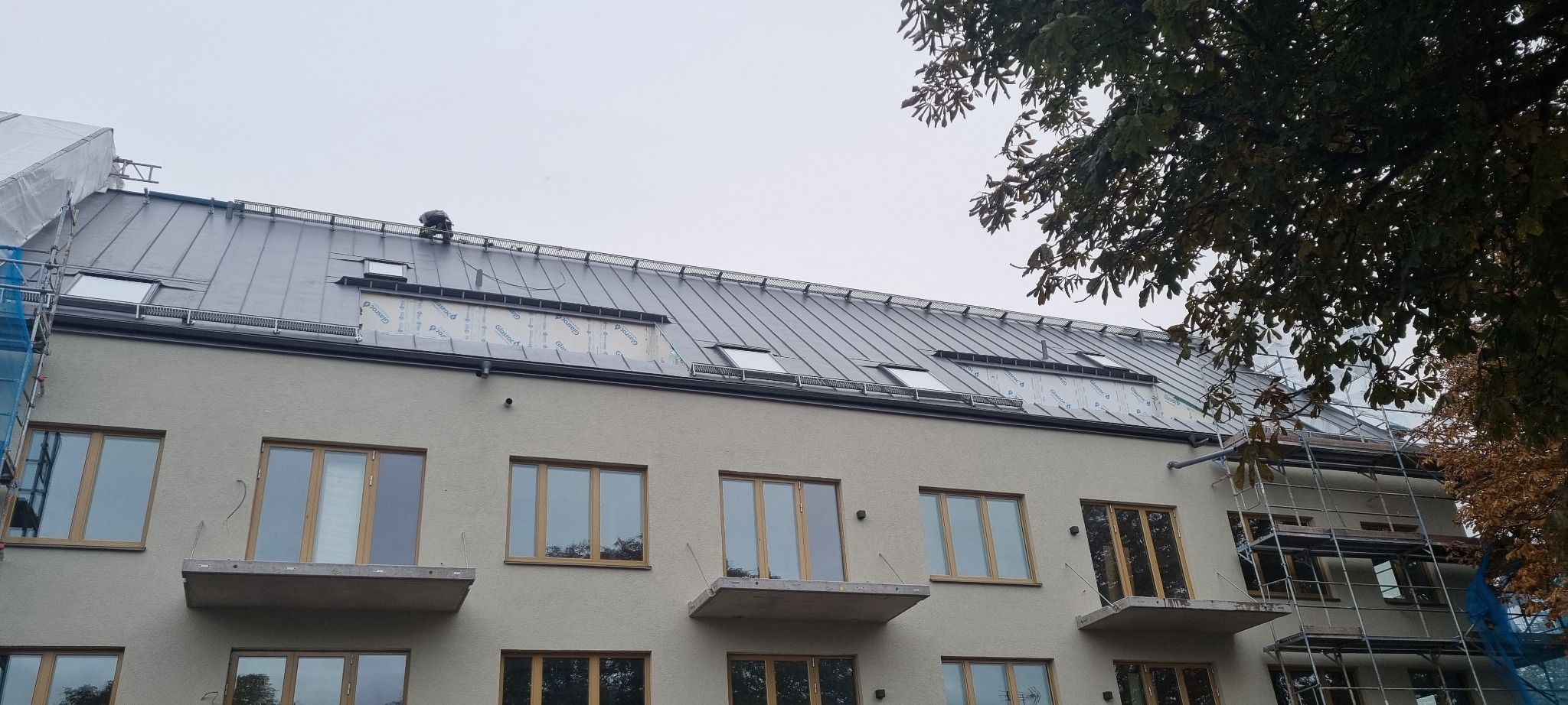 Montaż blachy na dachu budynku wielorodzinnego z oknami dachowymi, widoczne rusztowania i pracownik w odzieży roboczej.