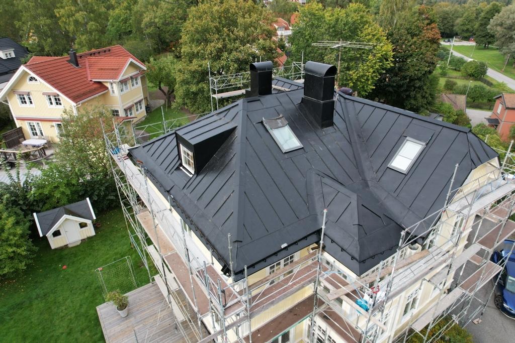 Czarny blaszany dach z oknami dachowymi i kominami, otoczony rusztowaniem, widok z góry na dom i ogród.