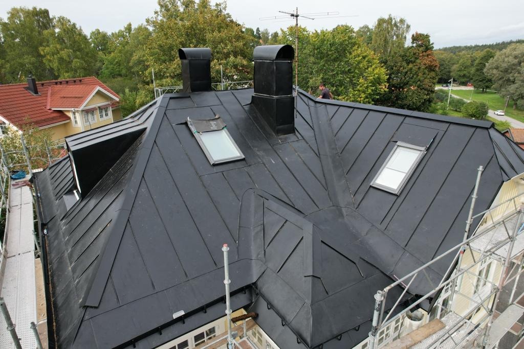 Czarny, metalowy dach z dwoma kominami i trzema oknami dachowymi, widziany z rusztowania. W tle drzewa i zabudowa jednorodzinna.