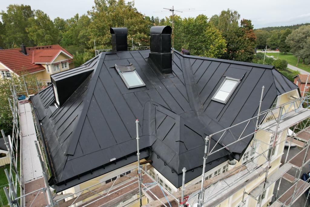 Czarny, metalowy dach z lukarnami i kominami, widziany z góry, z rusztowaniem w tle i zielenią drzew