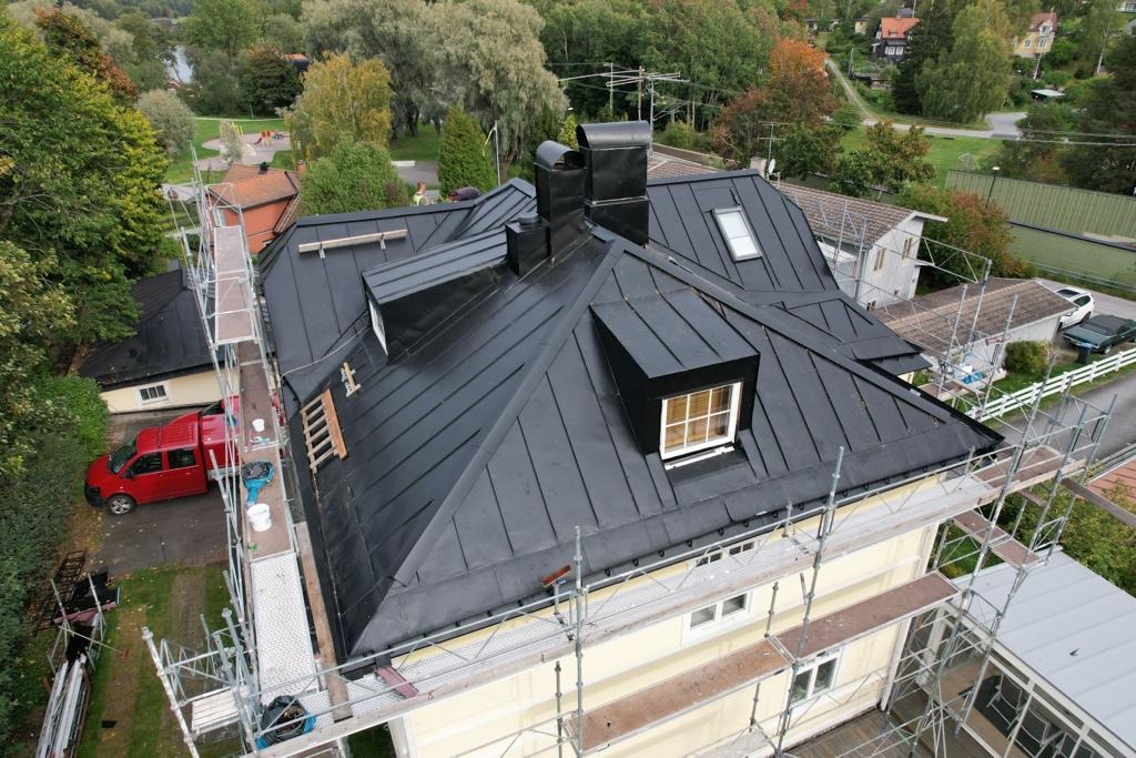 Czarny, metalowy dach z lukarnami i kominem, otoczony rusztowaniem, widok z góry na dom i okolicę z drzewami.