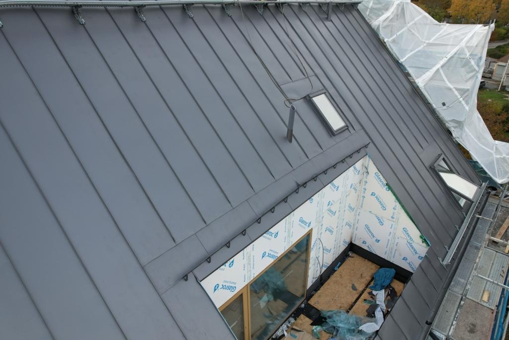 Szary dach z blachy na domu jednorodzinnym, z oknami dachowymi, widoczne prace wykończeniowe na poddaszu, zabezpieczone folią ściany, rusztowanie