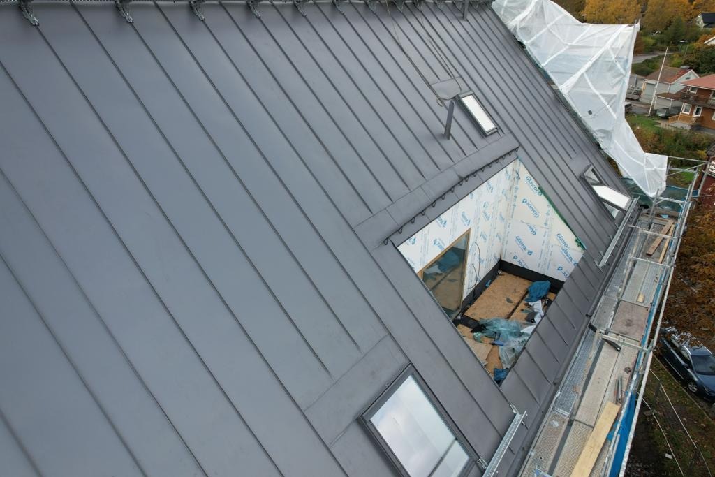 Szary dach z blachy na rąbek stojący, z oknami dachowymi i fragmentem poddasza w trakcie prac wykończeniowych, widoczne rusztowanie i zabezpieczenie folią.