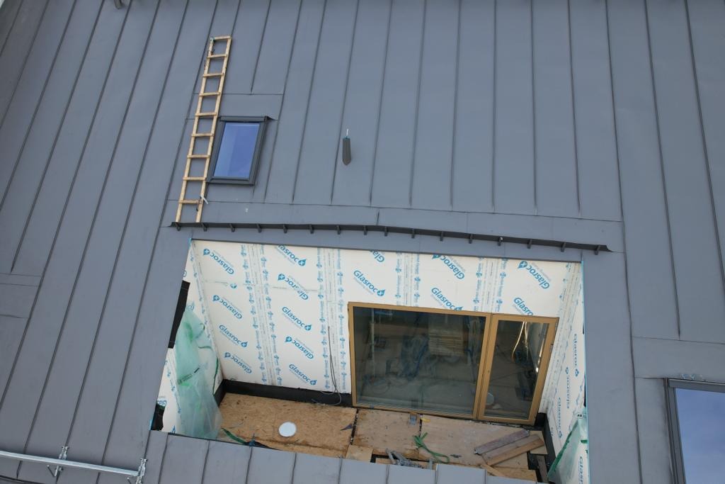 Szara blacha na dachu z oknem dachowym i drewnianą drabinką, widoczne prace przygotowawcze do montażu okien, ściany zabezpieczone folią Glasroc.