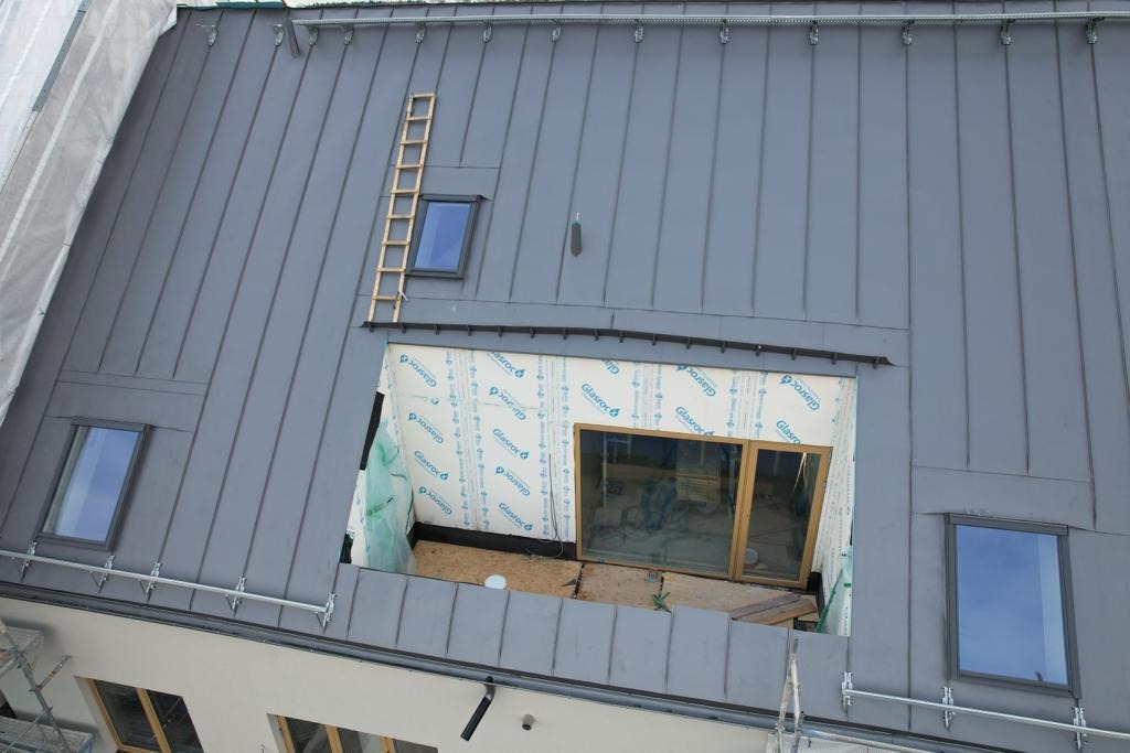 Szary dach z blachy na budynku w trakcie budowy, widoczne okna dachowe i otwór w dachu z materiałem izolacyjnym Glasroc, drabina oparta o dach.
