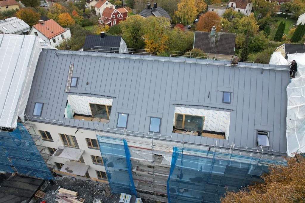 Wielorodzinny budynek z nowym, szarym dachem blaszanym z oknami dachowymi w trakcie montażu, widoczne rusztowania i pracownicy na dachu.