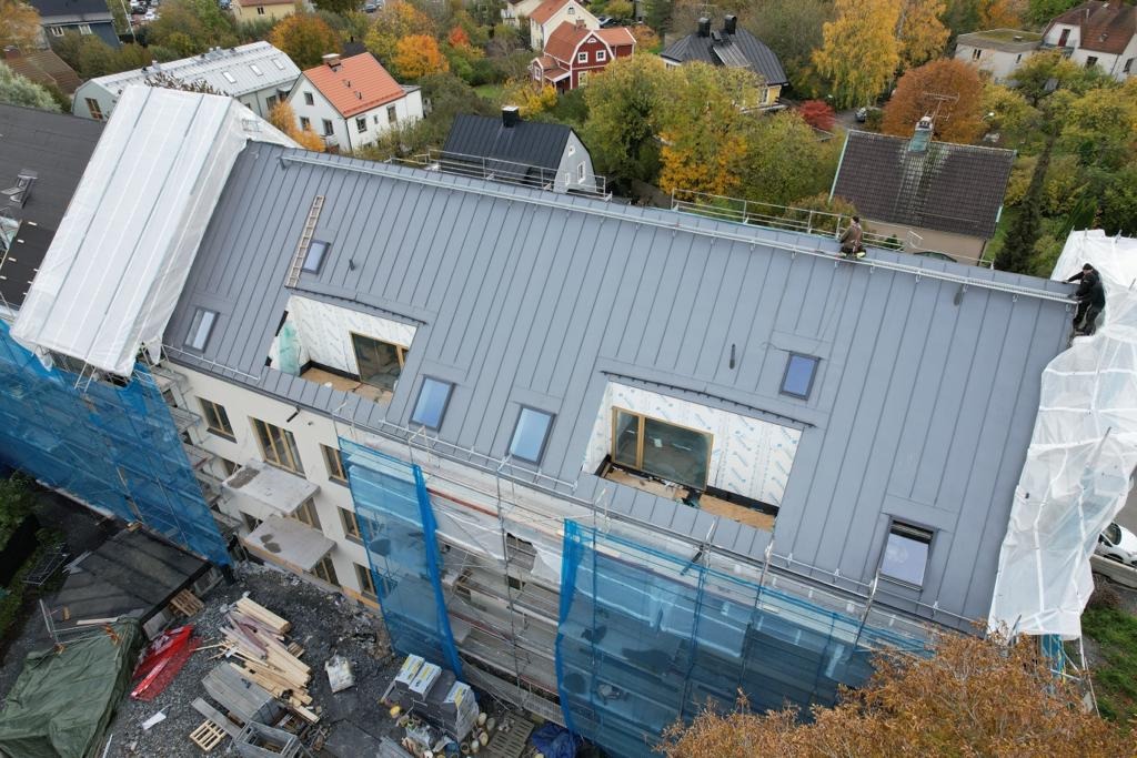 Wielorodzinny budynek w trakcie wymiany dachu z blachy na rąbek stojący; widoczne okna dachowe, zabezpieczenia rusztowaniami i materiały budowlane na placu budowy z perspektywy z góry.