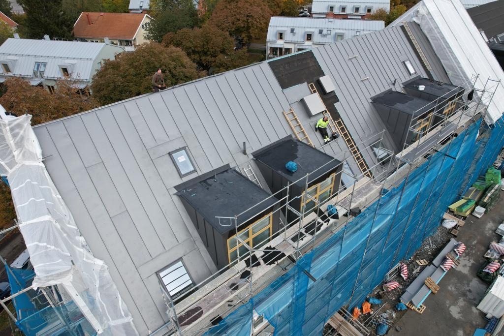 Widok z góry na dach budynku w trakcie prac remontowych, z widocznymi pracownikami, rusztowaniami zabezpieczonymi niebieską siatką, oknami dachowymi i elementami wykończeniowymi. Na dachu leżą...