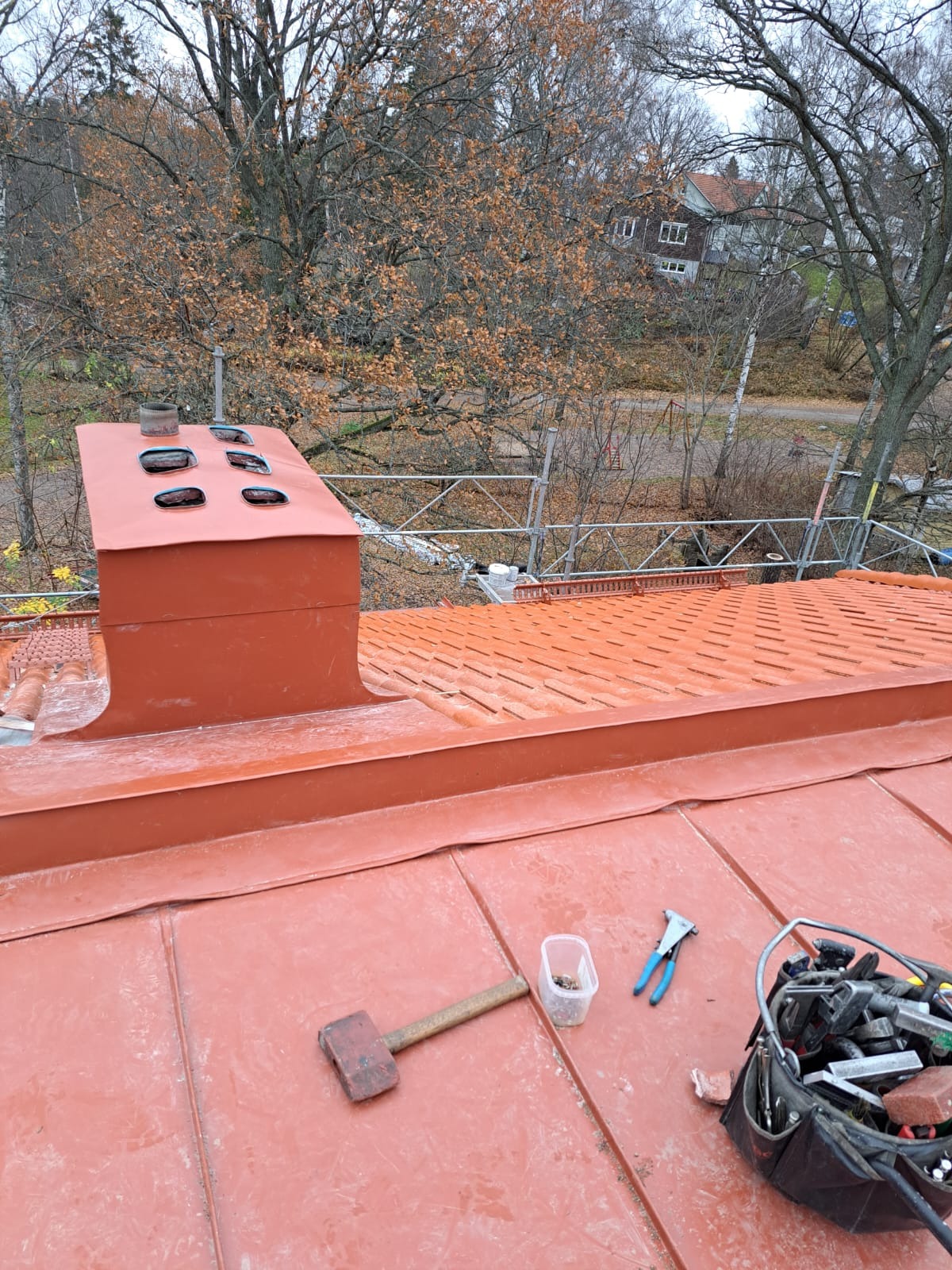 Czerwony dach z blachy trapezowej z kominem i narzędziami dekarskimi: młotek, nitownica, pojemnik na nity i torba z narzędziami. W tle drzewa bez liści i rusztowanie.