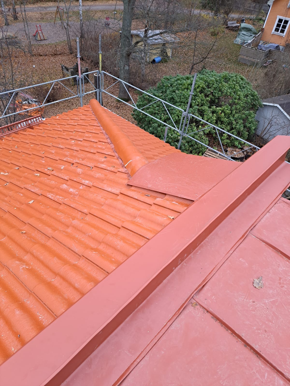 Czerwony dach z dachówki ceramicznej, częściowo pokryty płaskimi panelami, widoczny z rusztowania, z liśćmi na powierzchni i elementami wentylacyjnymi.