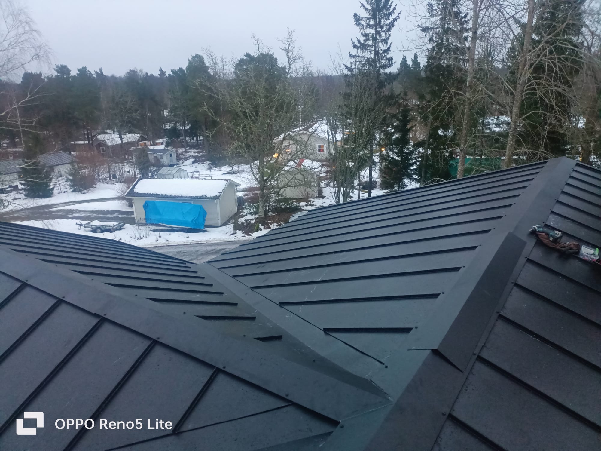 Nowy, ciemny dach z blachy na tle zimowego krajobrazu z ośnieżonymi budynkami i drzewami bez liści. Widoczne narzędzia dekarskie.