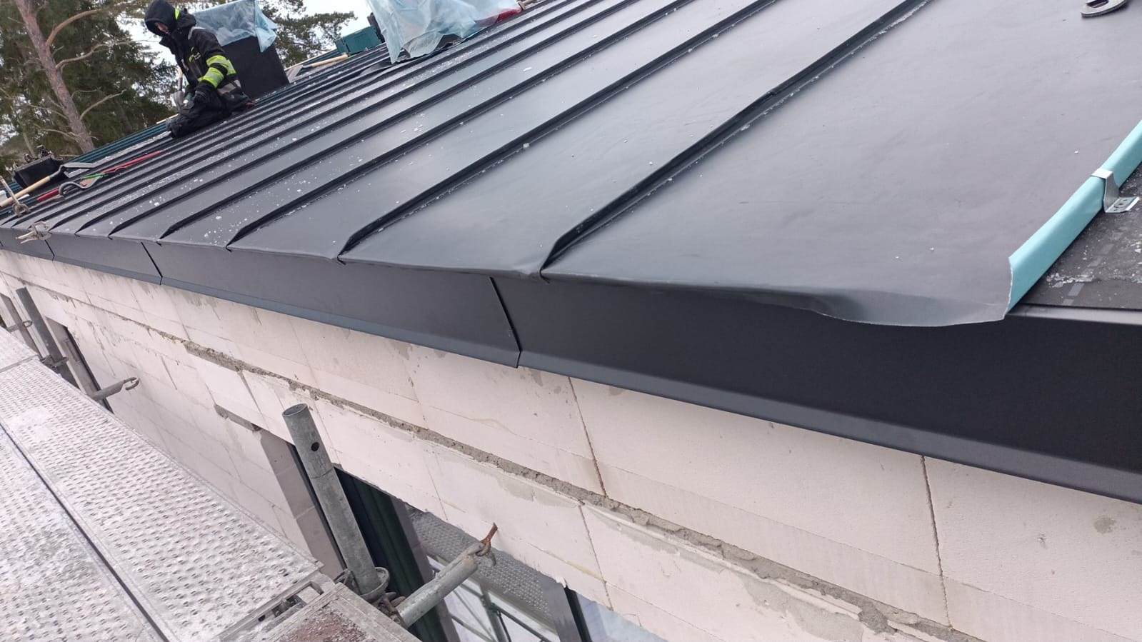 Montaż blachy na rąbek stojący na dachu budynku z białego bloczka, widoczny dekarz w kombinezonie na rusztowaniu.