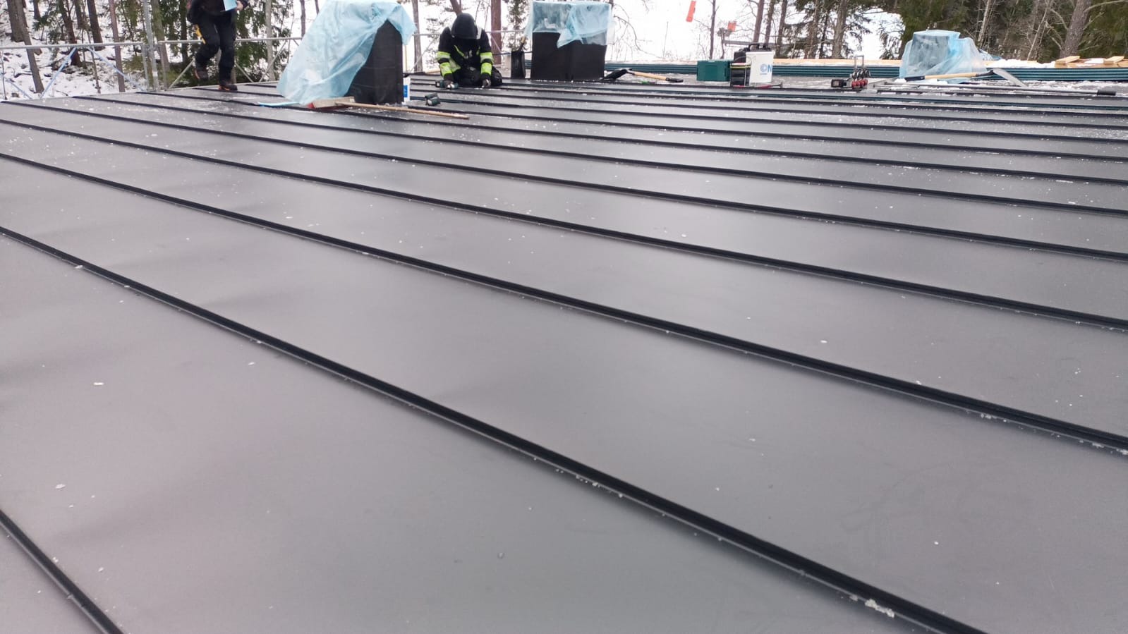 Montaż paneli na dachu z widocznymi pracownikami w odzieży roboczej i narzędziami, zimowy krajobraz w tle.