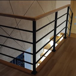 P. U. Stalowy Alex Michał Aleksanderek - Wnętrze domu z drewnianymi schodami i metalową balustradą z drewnianym poręczą, widok z góry na dół, jasna drewniana podłoga i drzwi.