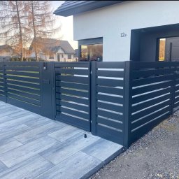 P. U. Stalowy Alex Michał Aleksanderek - Nowoczesna, antracytowa brama wjazdowa z poziomymi panelami na posesji z kostką brukową i domem w tle. Widoczny czarny pojemnik na bioodpady.