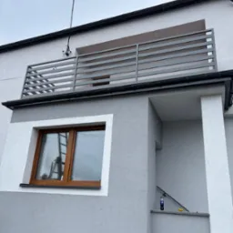 Nowoczesna, pozioma balustrada balkonowa z szarych profili na tle szarej elewacji domu. Widok z dołu, okno z drewnianą ramą, fragment schod&oacute;w.