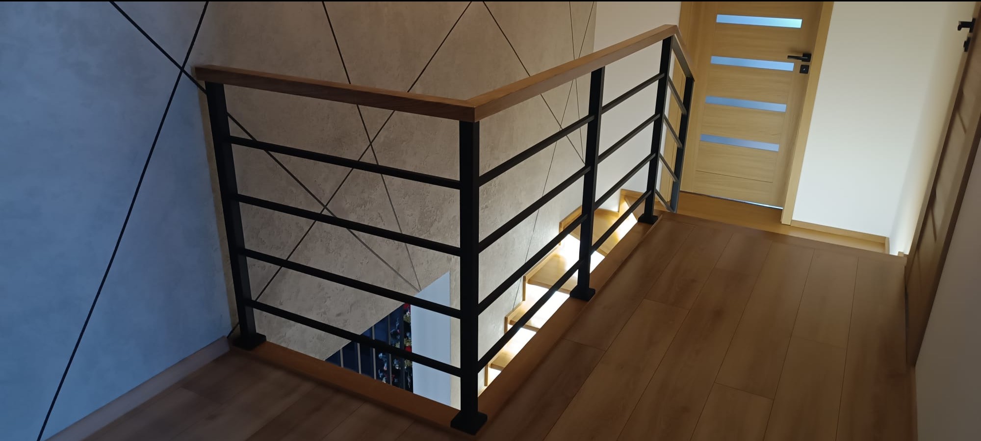 Wnętrze domu z drewnianymi schodami i metalową balustradą z drewnianym poręczą, widok z góry na dół, jasna drewniana podłoga i drzwi.