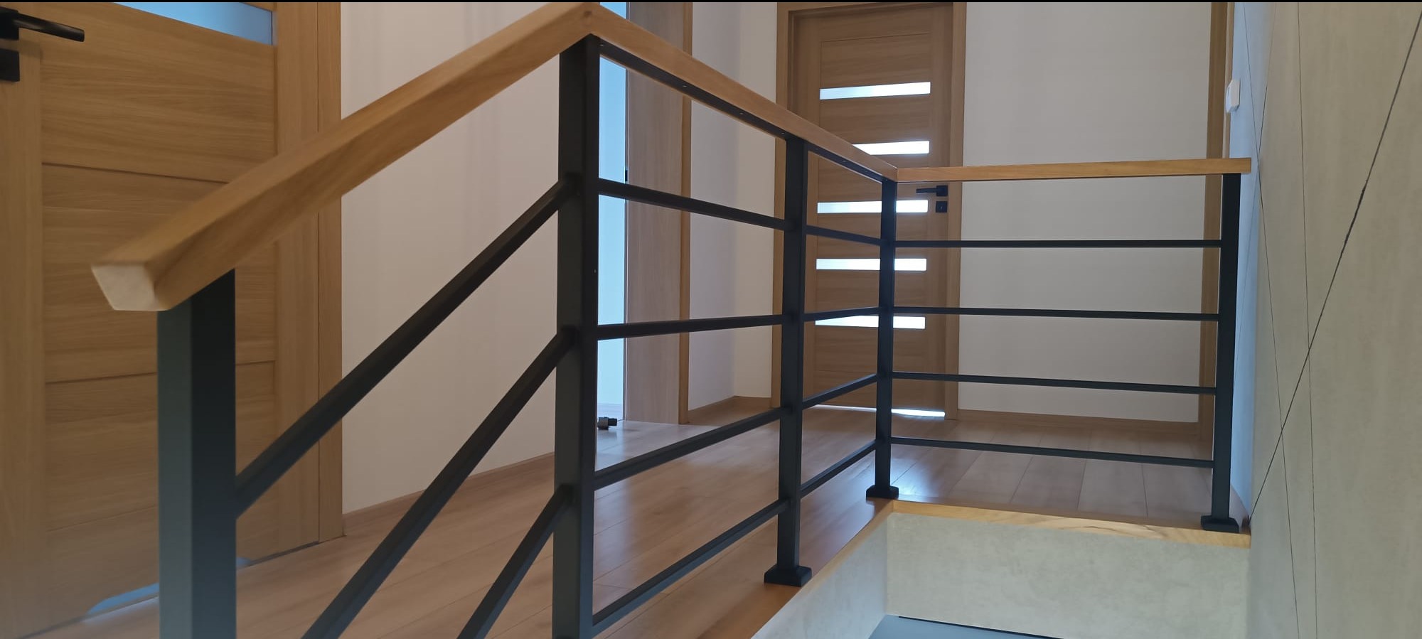 Nowoczesna balustrada schodowa z czarnych metalowych prętów i drewnianym pochwytem, widok z góry na jasne drewniane schody i drzwi.