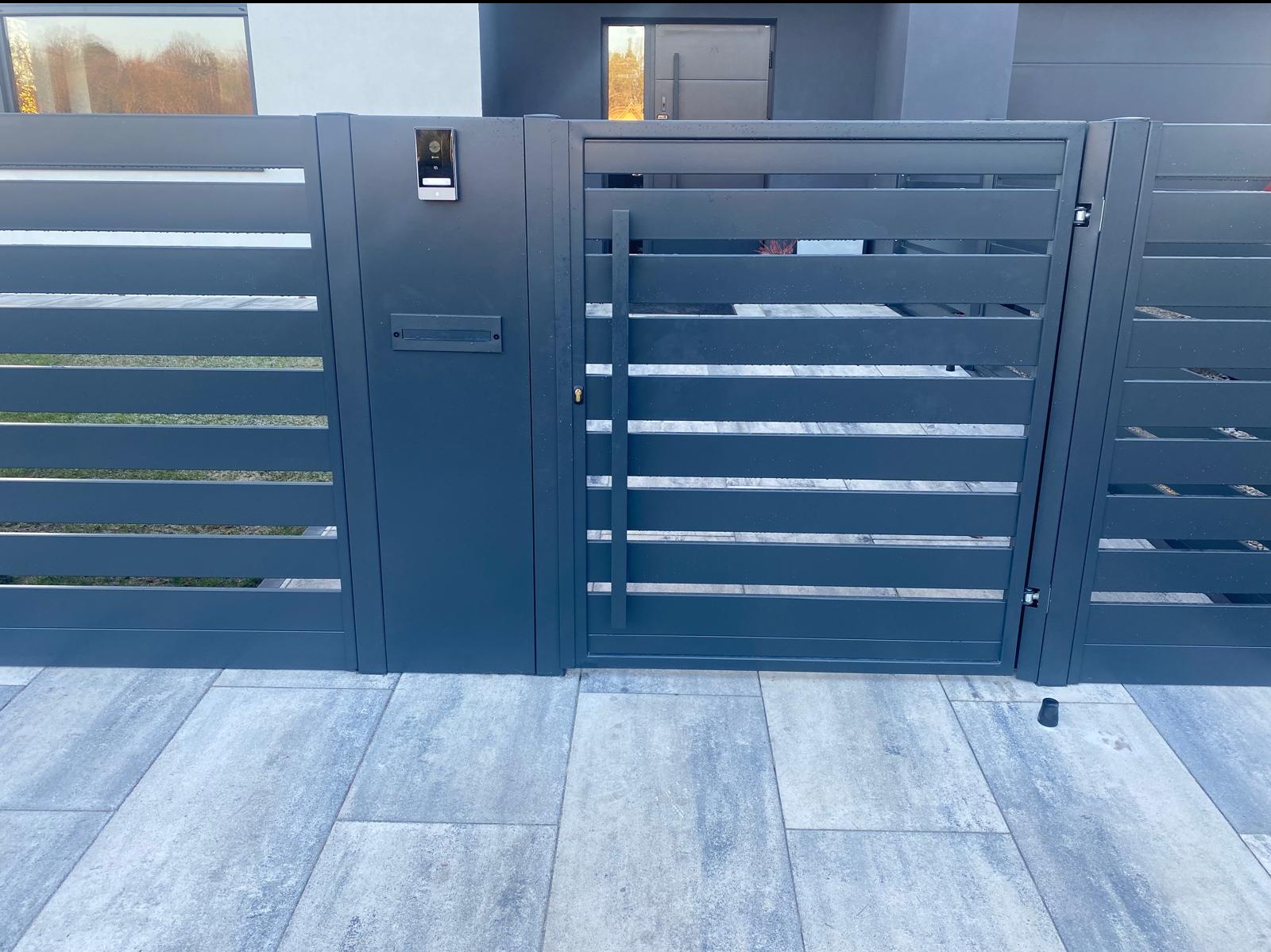 Nowoczesna brama wjazdowa z poziomymi panelami w kolorze grafitowym, z wideodomofonem i skrzynką na listy, na tle szarej kostki brukowej.