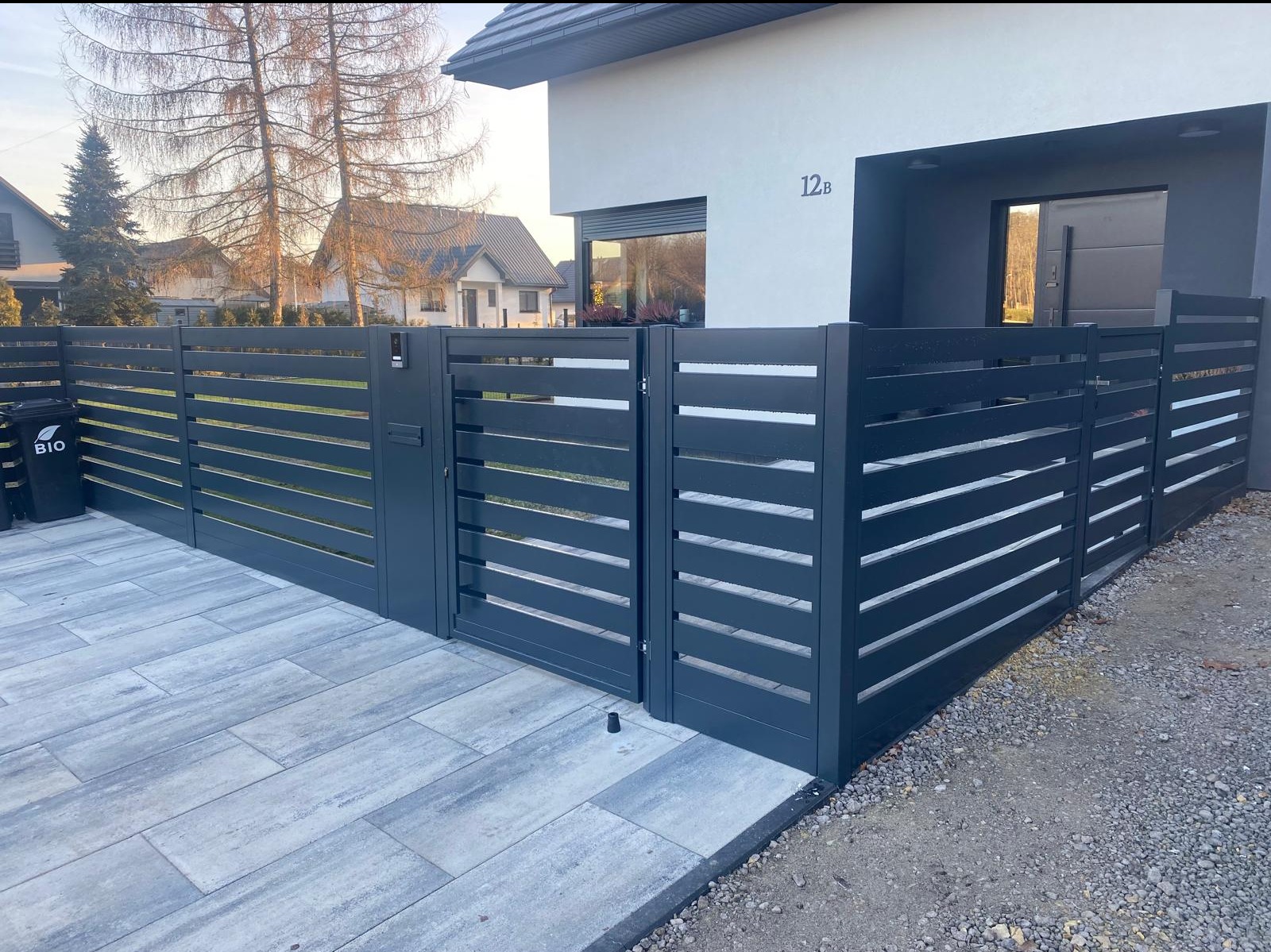 Nowoczesna, antracytowa brama wjazdowa z poziomymi panelami na posesji z kostką brukową i domem w tle. Widoczny czarny pojemnik na bioodpady.