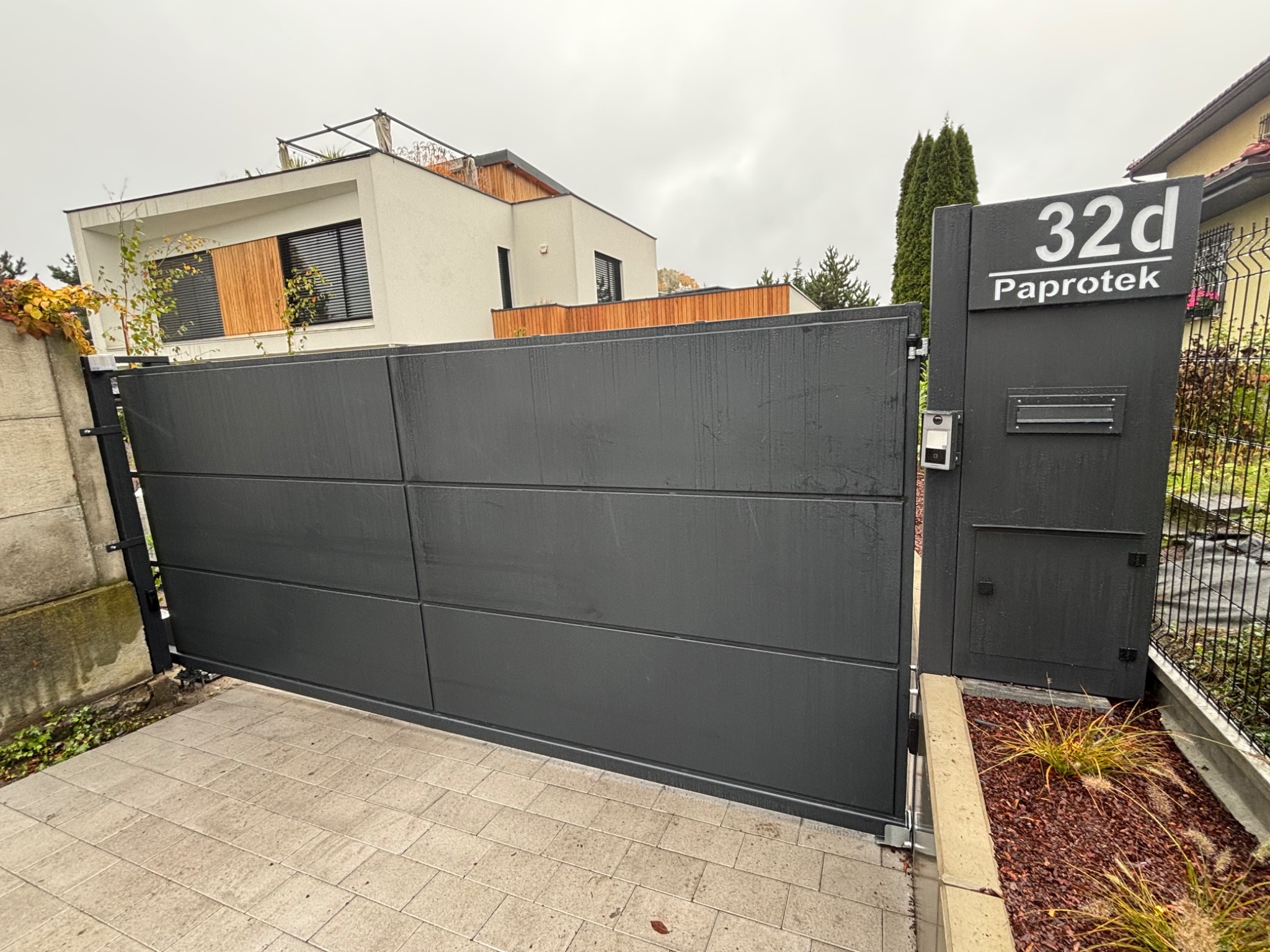 Nowoczesna, przesuwna brama wjazdowa w kolorze grafitowym, z numerem domu '32d Paprotek' na słupku. Brama zintegrowana ze skrzynką pocztową i domofonem.