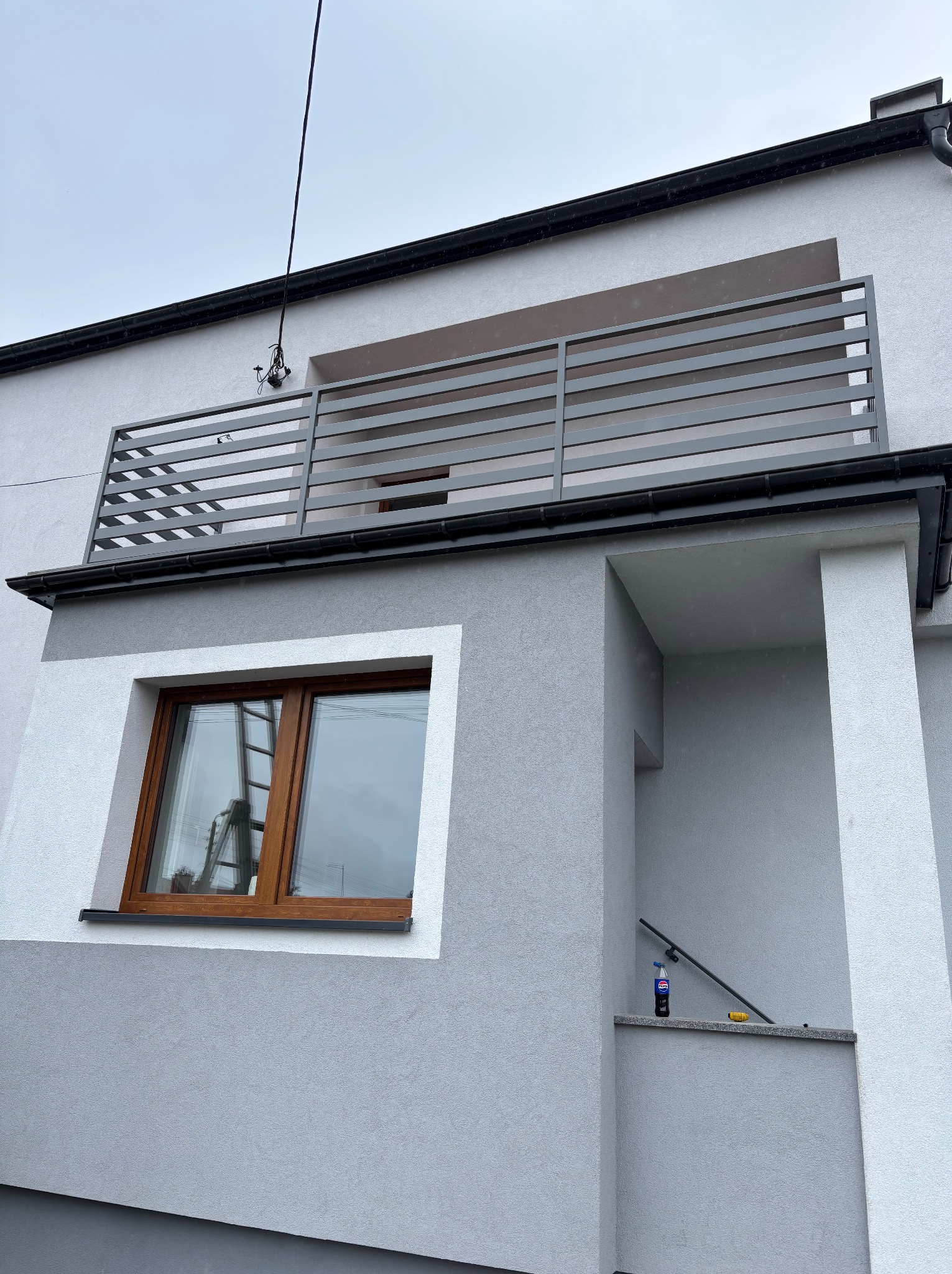 Nowoczesna, pozioma balustrada balkonowa z szarych profili na tle szarej elewacji domu. Widok z dołu, okno z drewnianą ramą, fragment schodów.