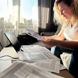 Kobieta analizuje dokumenty przy laptopie w domowym biurze. Na stole rozłożone arkusze z tabelami, w tle okno i zasłony. Słońce oświetla dokumenty.