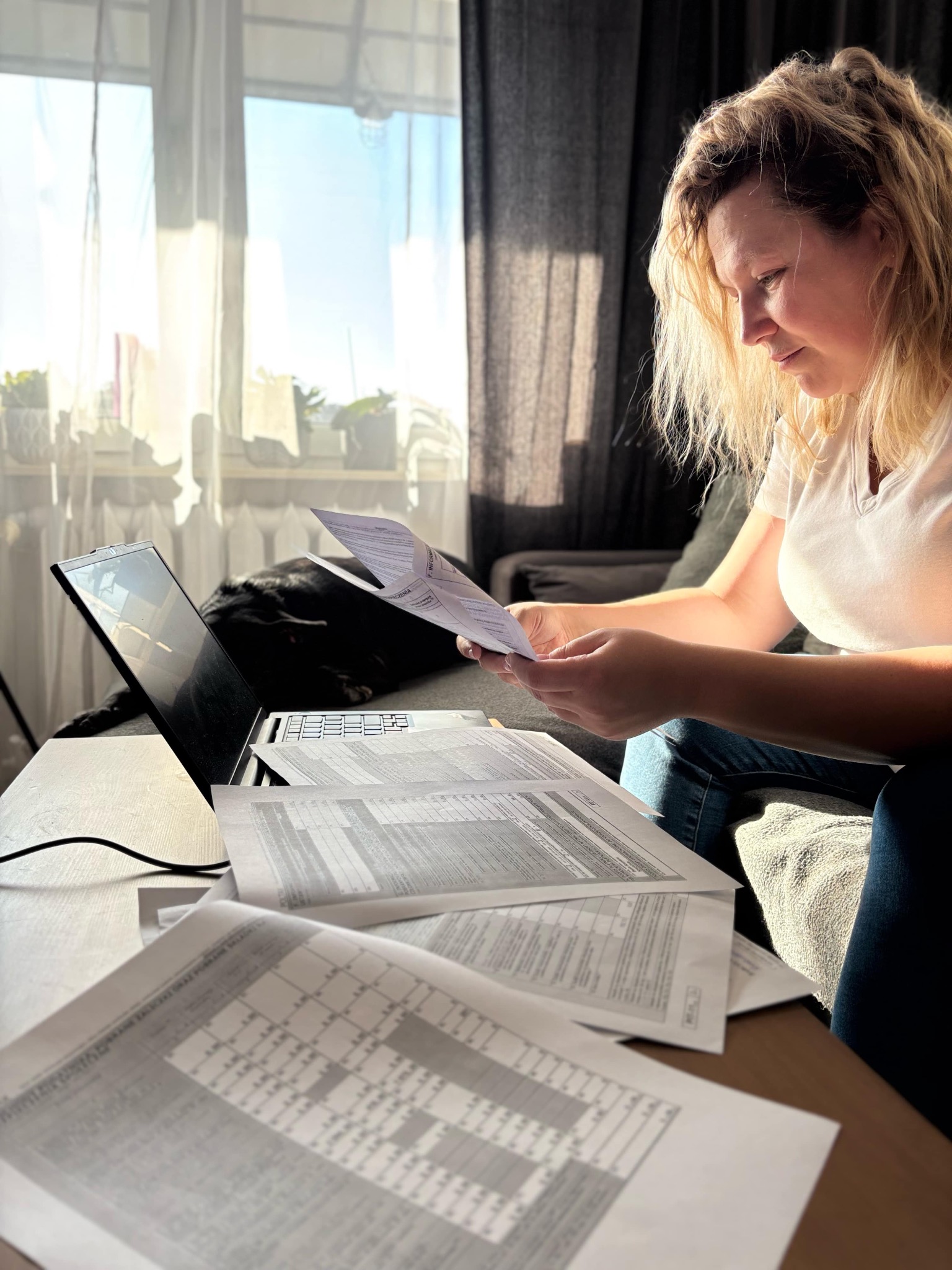 Kobieta analizuje dokumenty przy laptopie w domowym biurze. Na stole rozłożone arkusze z tabelami, w tle okno i zasłony. Słońce oświetla dokumenty.