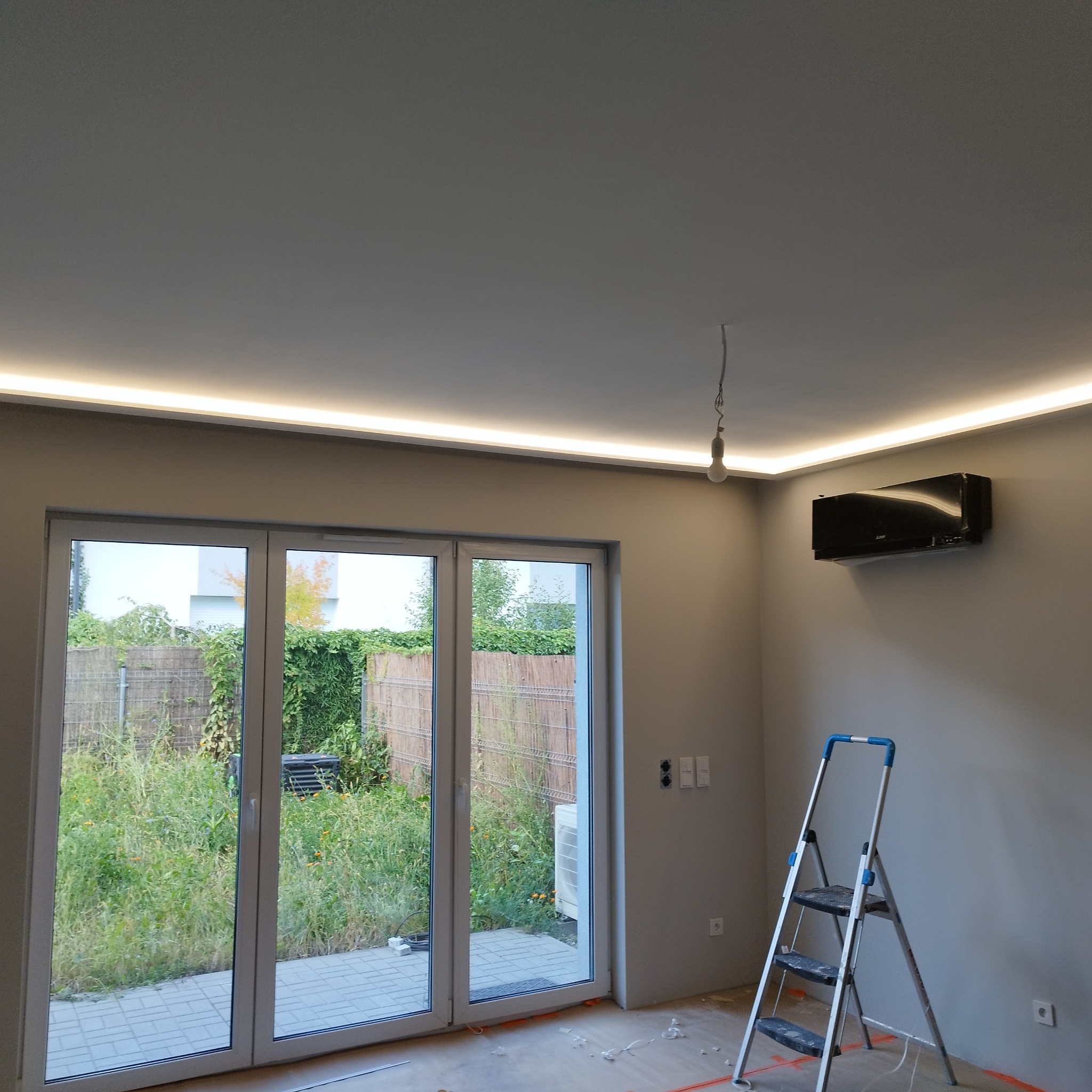 Wnętrze w trakcie prac wykończeniowych: zamontowany klimatyzator, listwa LED pod sufitem, widoczne ślady po remoncie na podłodze, drabina aluminiowa, przesuwne drzwi balkonowe z widokiem...