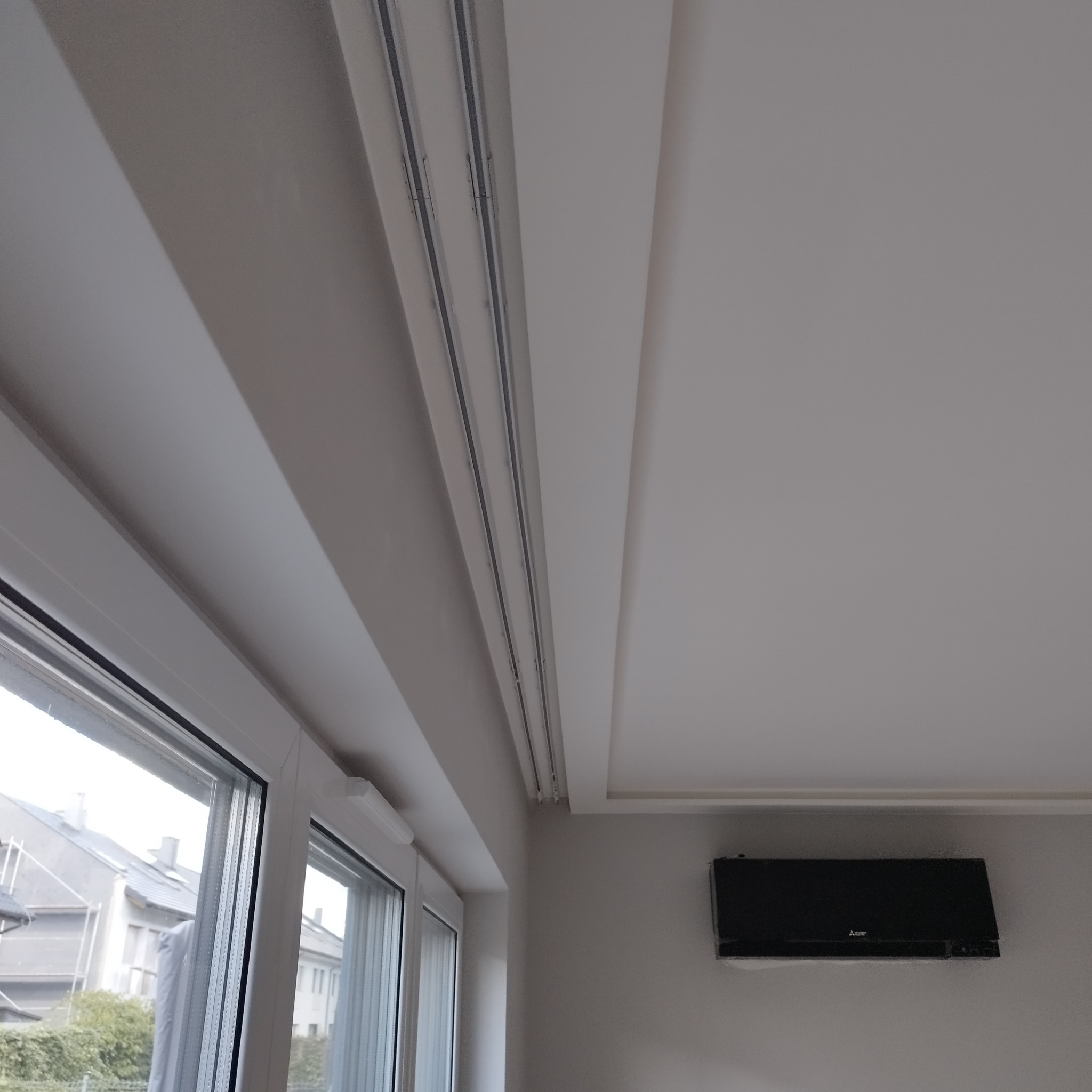 Szare wnętrze pokoju po remoncie z widocznymi szynami karnisza podsufitowego, oknem z żaluzjami i zamontowanym klimatyzatorem.