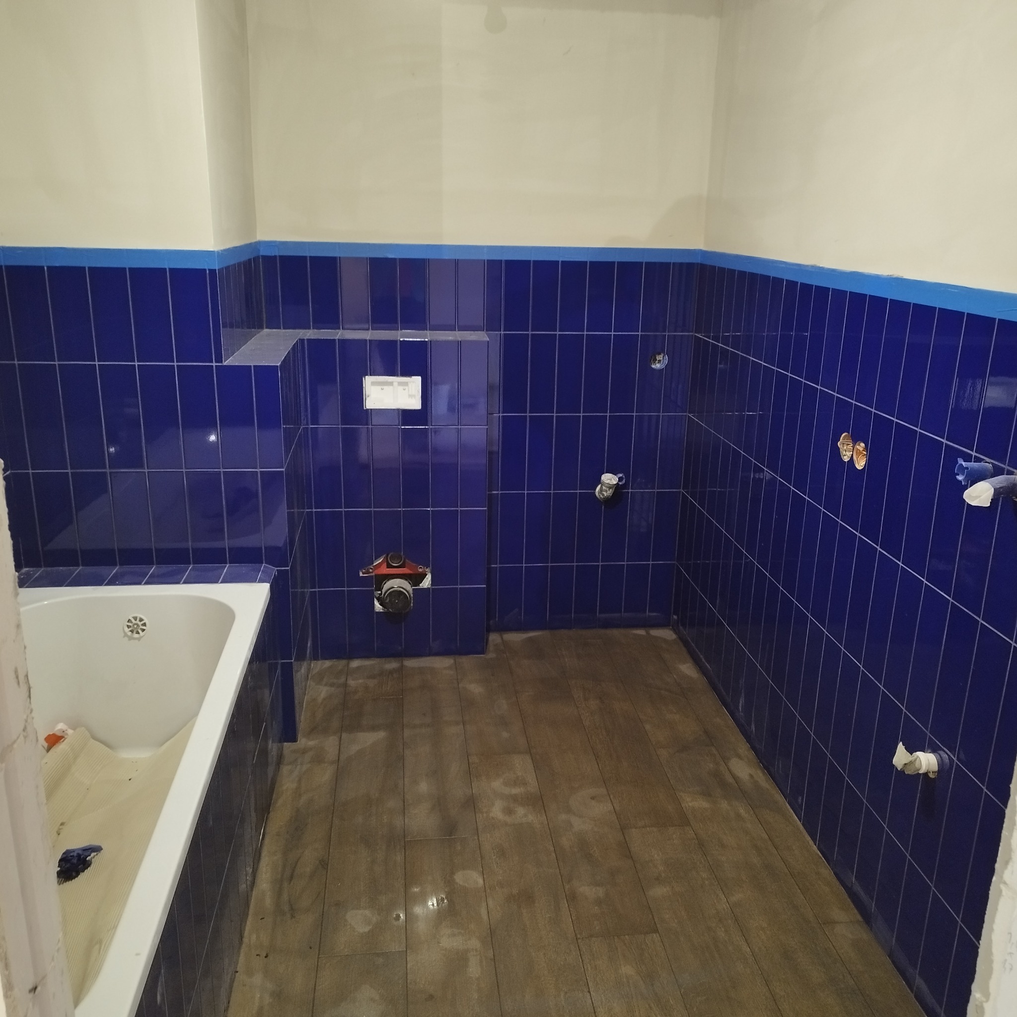 Niebieskie, pionowo ułożone płytki w trakcie układania w łazience, widoczne fragmenty instalacji hydraulicznej i przygotowana wanna z zabezpieczeniem, podłoga wyłożona panelami imitującymi drewno.