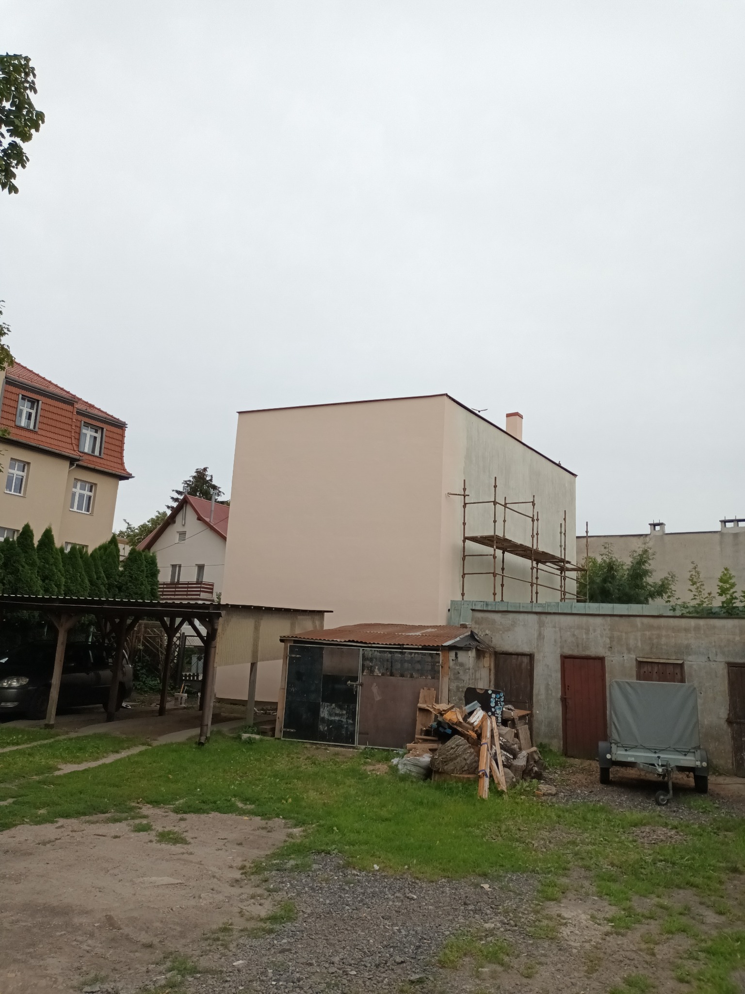 Budynek po odświeżeniu elewacji, widoczne rusztowania, na dole podwórko z przyczepką i stertą drewna.