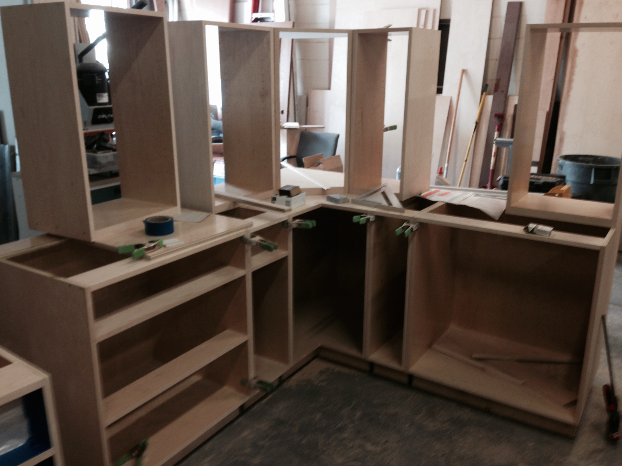 Fragment montażu drewnianych mebli kuchennych w warsztacie, widoczne klamry, śruby i narzędzia, surowe drewno i elementy konstrukcyjne.