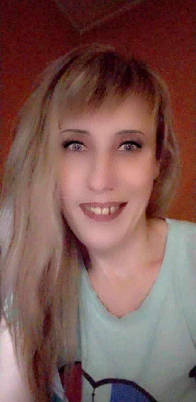 Portret kobiety z blond włosami i jasną cerą, uśmiechającej się do kamery na rozmytym tle.