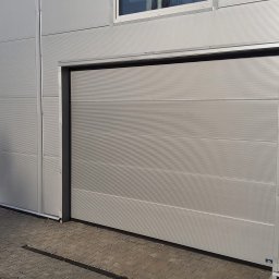 Stan-Mal R. Malicki Sp.j - Zamknięta, pozioma brama garażowa w kolorze szarym, z delikatnym wzorem prążków, zamontowana w budynku o metalowej elewacji. Widoczny fragment chodnika z kostki brukowej.