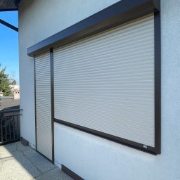 Stan-Mal R. Malicki Sp.j - Zamknięta roleta zewnętrzna w brązowej obudowie, zamontowana na elewacji budynku. Widoczny fragment balkonu i niebieskie niebo w tle.