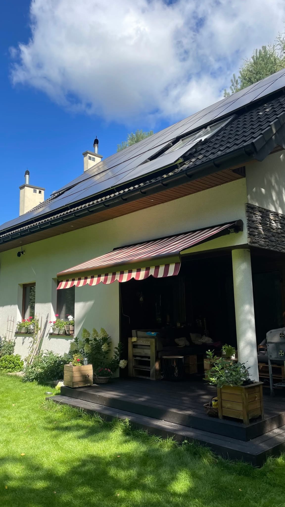 Elewacja domu z markizą w biało-czerwone pasy nad tarasem. Na dachu panele słoneczne, okna z kwiatami. Widok z ogrodu w słoneczny dzień.