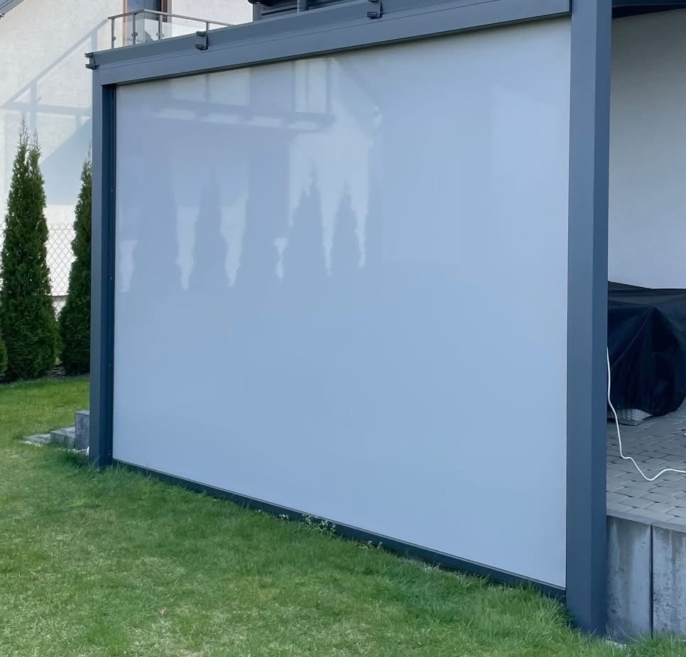 Szara, zwijana osłona przeciwsłoneczna zamontowana na konstrukcji pergoli, widoczna na tle zielonego trawnika i fragmentu budynku.