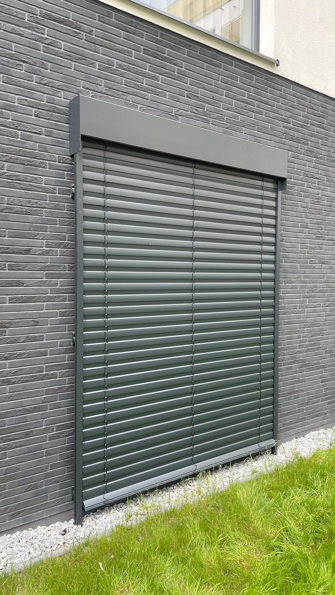 Szara, zamknięta żaluzja fasadowa na tle ściany z szarej cegły, z widoczną zielenią trawy u dołu. Minimalistyczny design, równoległe linie, nowoczesna architektura.
