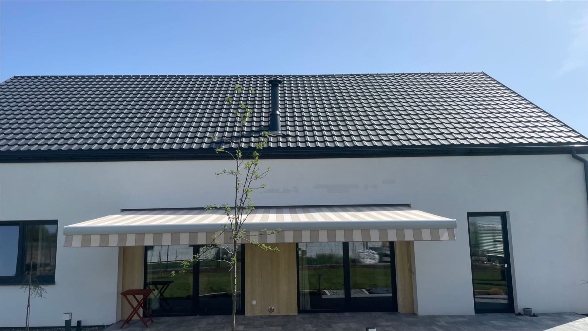 Elegancka markiza w beżowo-białe pasy nad tarasem domu z szarą dachówką. Widoczny fragment ogrodu i stolik. Jasne tło budynku i błękitne niebo.