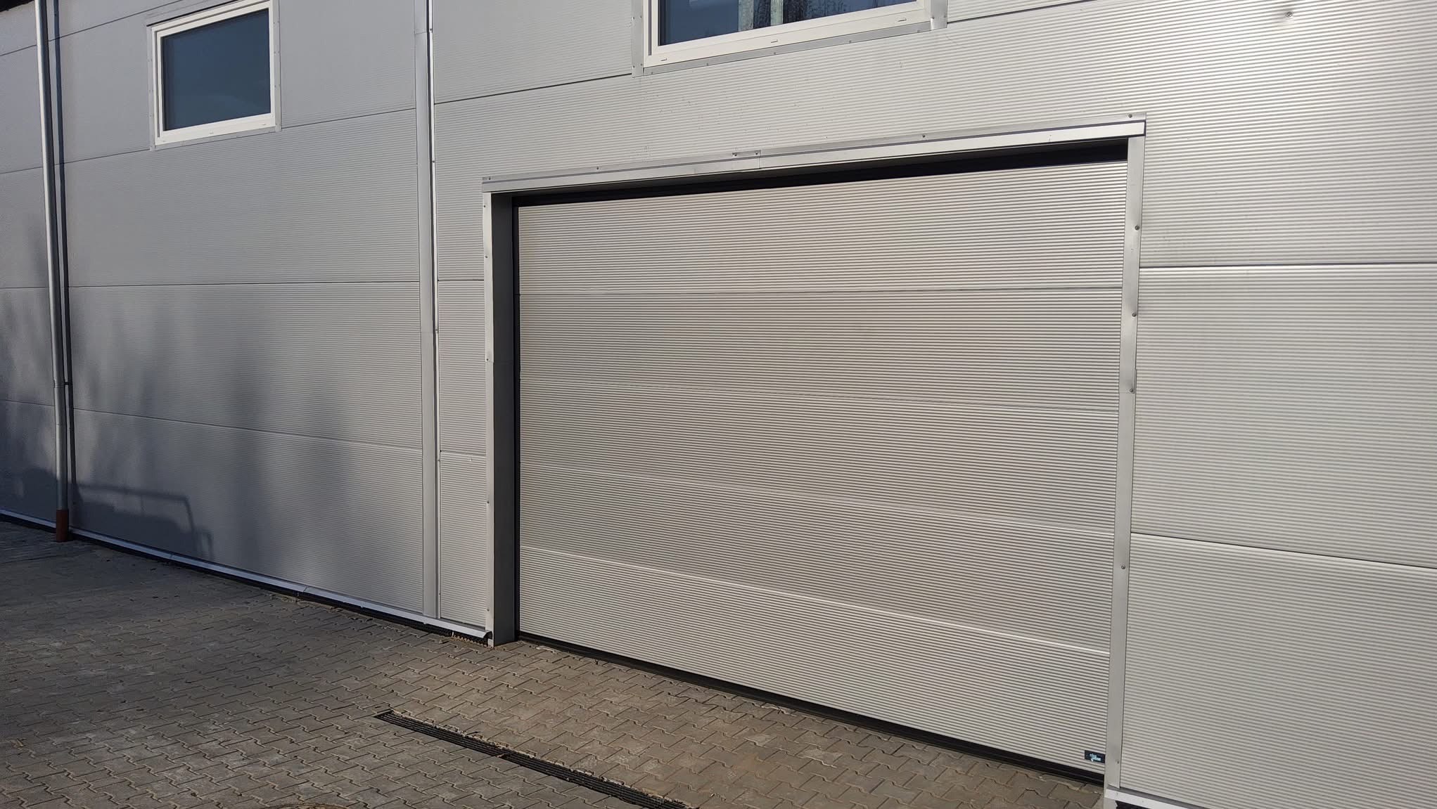 Zamknięta, pozioma brama garażowa w kolorze szarym, z delikatnym wzorem prążków, zamontowana w budynku o metalowej elewacji. Widoczny fragment chodnika z kostki brukowej.
