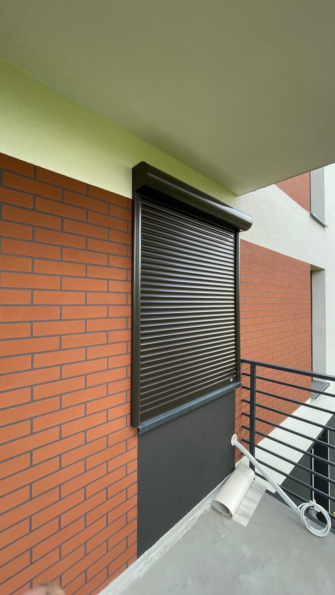 Ciemnobrązowa roleta zewnętrzna, zamontowana na balkonie budynku z ceglaną elewacją. Widoczne elementy montażowe i fragment balkonu z barierką.