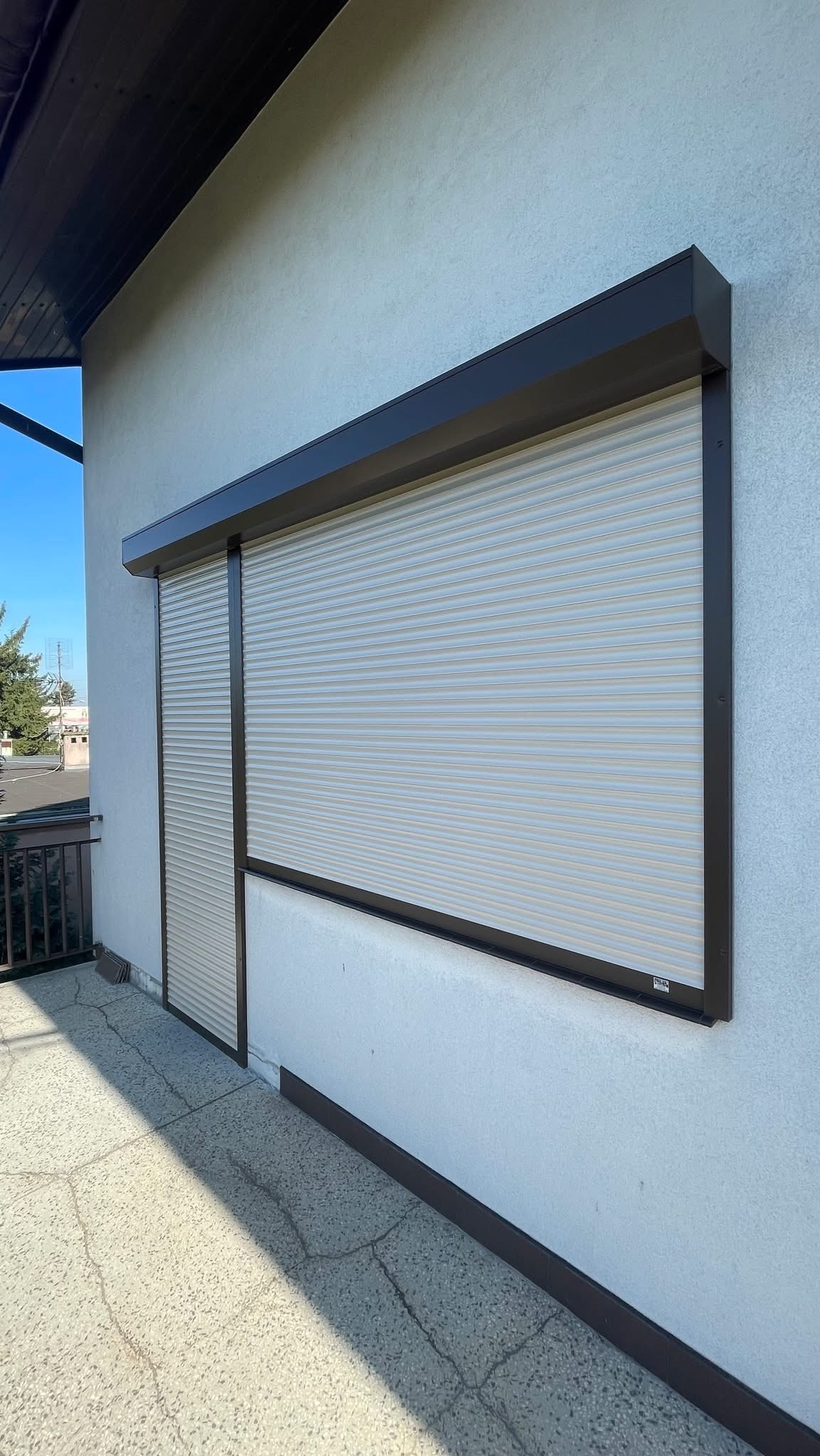 Zamknięta roleta zewnętrzna w brązowej obudowie, zamontowana na elewacji budynku. Widoczny fragment balkonu i niebieskie niebo w tle.