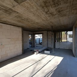 Nest House Development Dawid Fijałkowski - Wnętrze surowego domu w trakcie budowy z widocznymi ścianami z bloczków, betonowym stropem i podporami. Na posadzce leżą narzędzia i materiały budowlane, a przez otwory okienne wpada światło.