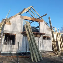 Budowa domu murowanego w stanie surowym otwartym, widoczne bloczki, elementy konstrukcji dachu, drewniane podpory i deski, drugi dom w tle.