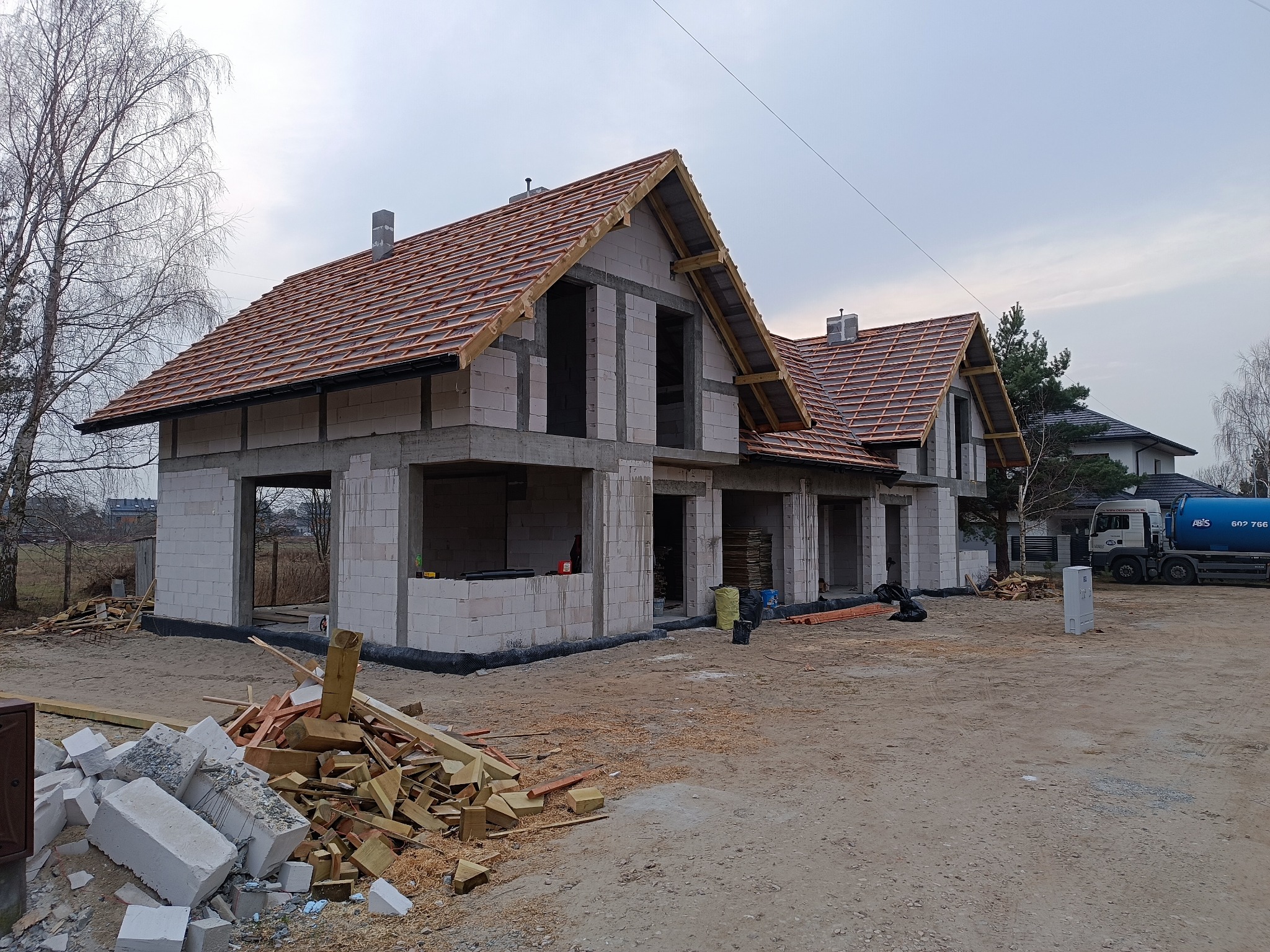 Surowy stan domu z białego bloczka, widoczna konstrukcja dachu pokryta dachówką, na pierwszym planie sterta gruzu i drewna, w tle zaparkowany cysterna z wodą.