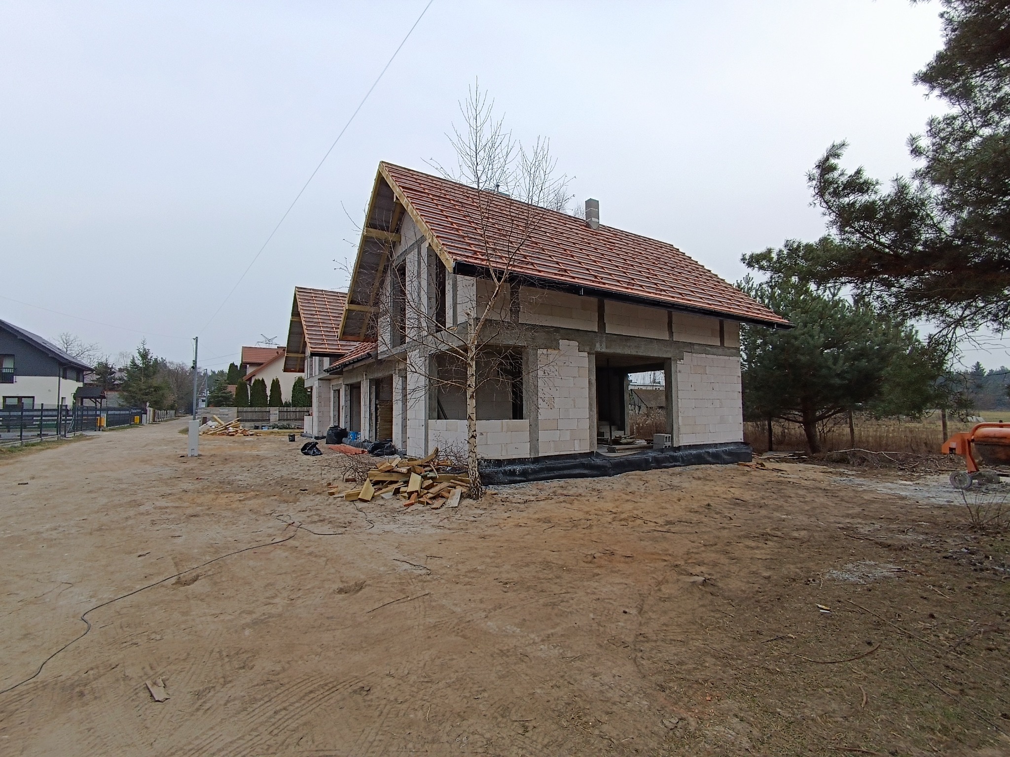 Surowy stan budynku jednorodzinnego z czerwonym dachem, widoczne białe bloczki konstrukcyjne i elementy drewniane więźby dachowej, plac budowy z ziemią i resztkami drewna.