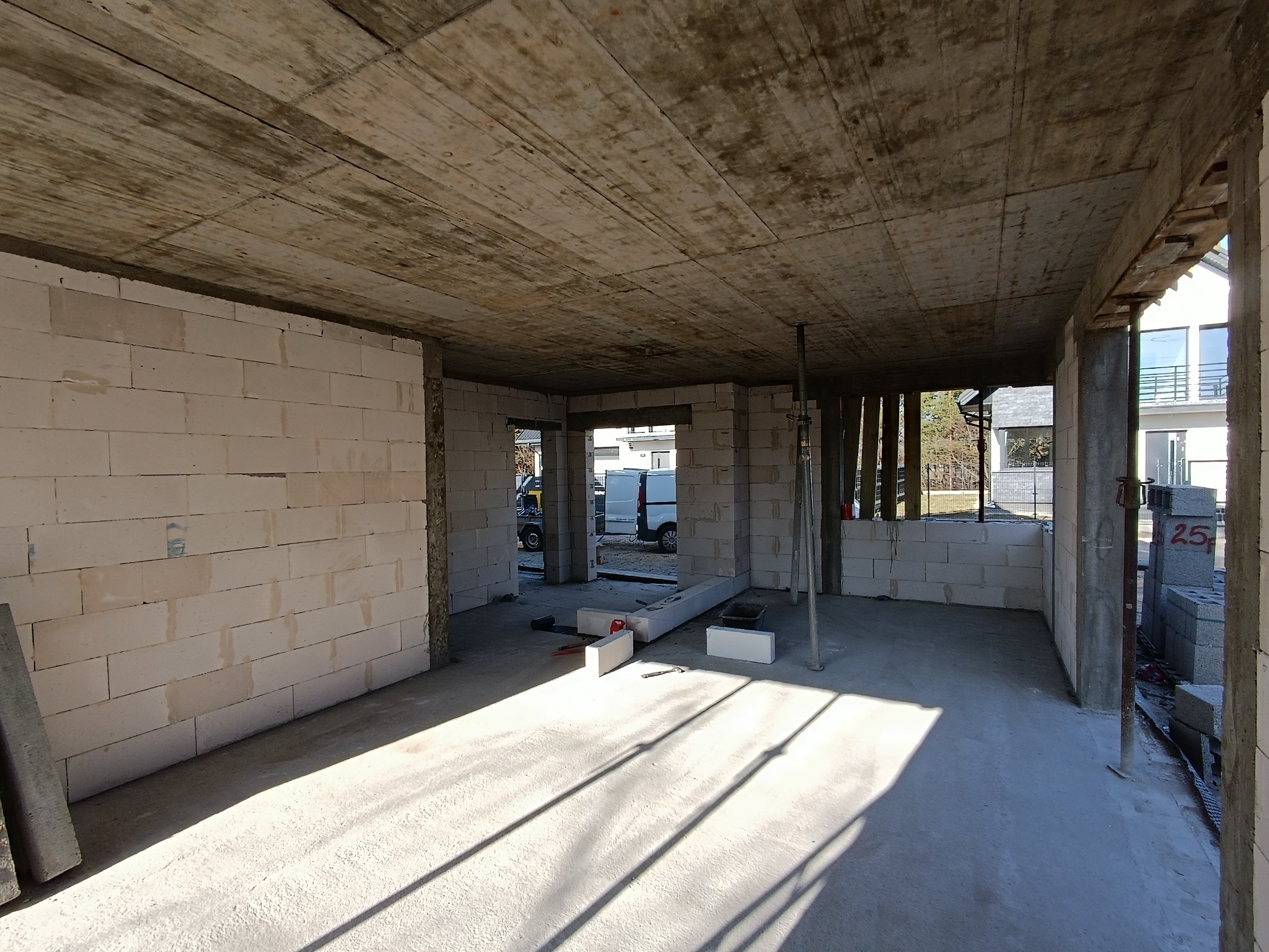 Wnętrze surowego domu w trakcie budowy z widocznymi ścianami z bloczków, betonowym stropem i podporami. Na posadzce leżą narzędzia i materiały budowlane, a przez otwory okienne wpada światło.