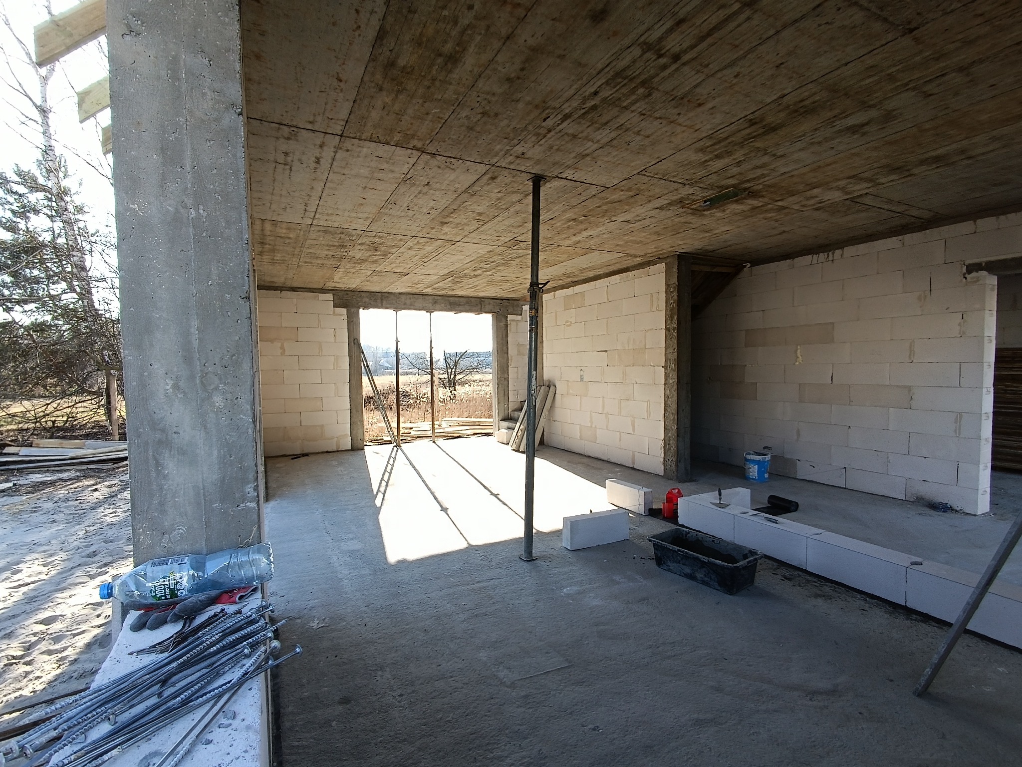 Surowy stan wnętrza domu murowanego z widoczną konstrukcją betonową stropu podpartą metalową podporą, ściany z bloczków, narzędzia i materiały budowlane wewnątrz, wpadające światło słoneczne.